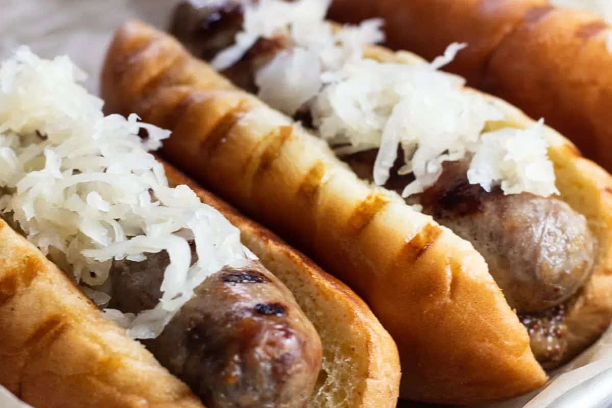 Three grilled sausages in buns topped with sauerkraut are arranged close together on a plate, making them perfect for Oktoberfest recipes or anyone craving easy German food.
