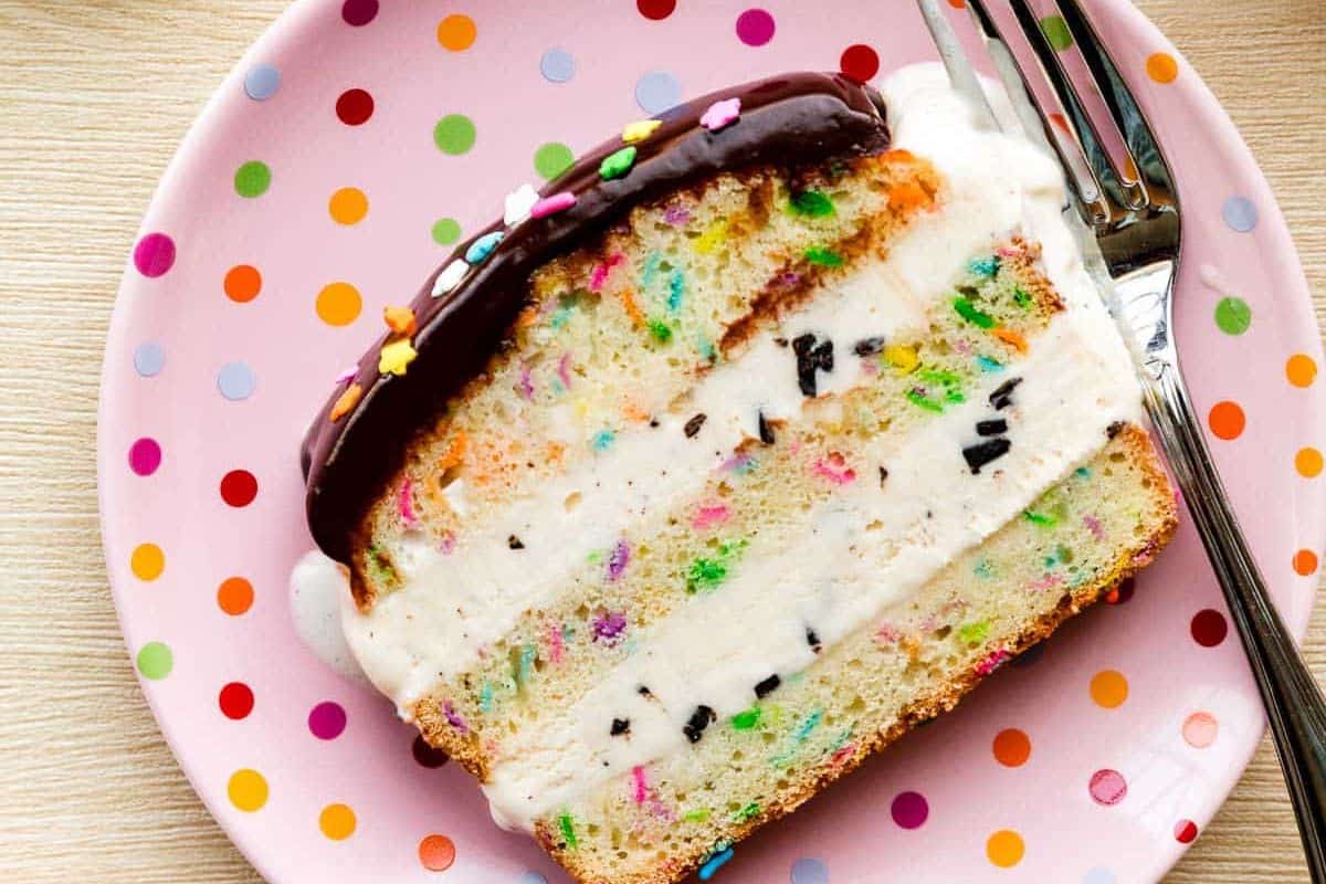 A slice of ice cream cake with colorful sprinkles and chocolate topping on a pink polka dot plate, next to a fork—a fun pick for easy dessert recipes or wow desserts at any summer gathering.