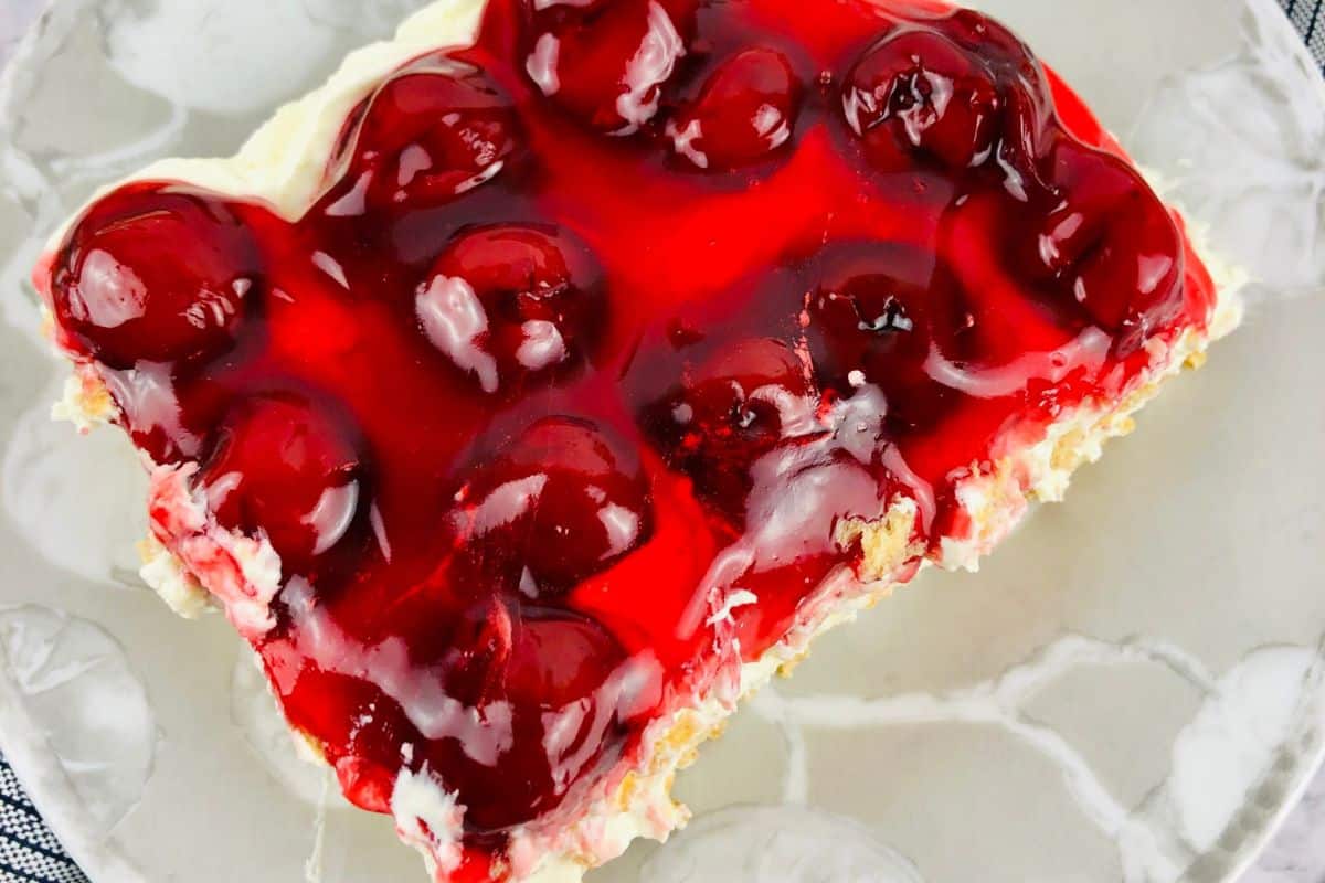 A slice of cherry dessert from our Fresh Cherry Recipes topped with whole cherries in red gelatin on a cream-filled crust, served on a clear glass plate.