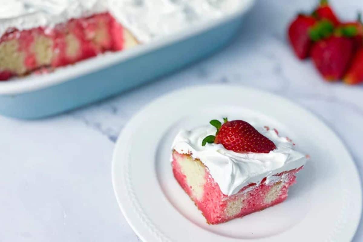 A slice of strawberry poke cake topped with whipped cream and a strawberry sits on a white plate, with the rest of the cake and fresh strawberries in the background—one of those wow desserts perfect for summer dessert recipes.