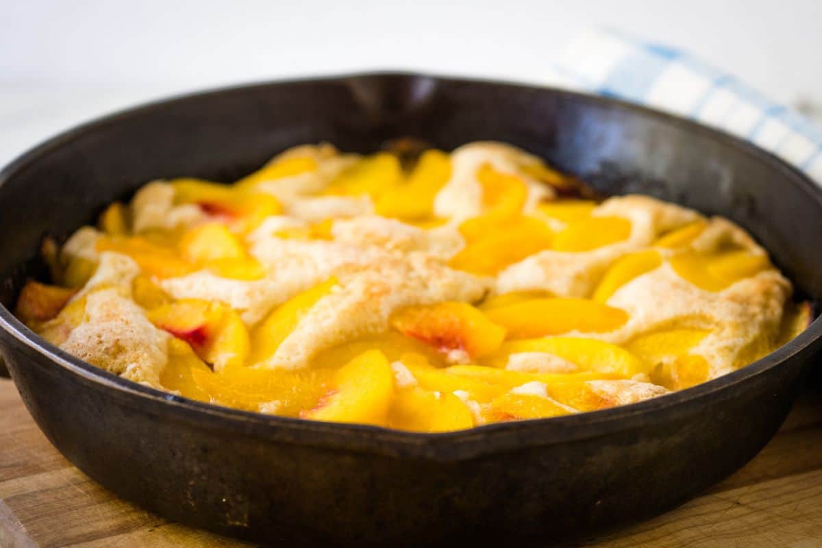 A peach cobbler with golden crust and sliced peaches, baked in a cast iron skillet, sits on a wooden surface—one of those wow dessert recipes perfect for any gathering.