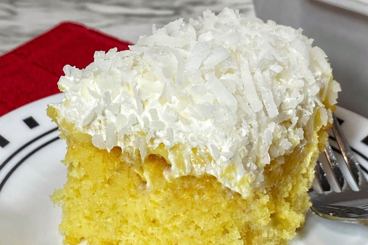 A square slice of yellow cake with white frosting and shredded coconut on top, served on a white plate with a fork—perfect for fans of irresistible coconut recipes.