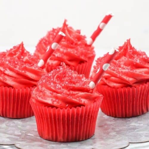 Four red frosted cupcakes with red sprinkles and polka dot straws are arranged on a scalloped metal tray—perfect for fans of cherry desserts or anyone searching for fresh cherry recipes.