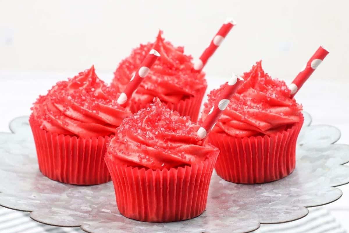 Four red frosted cupcakes with red sprinkles and polka dot straws are arranged on a scalloped metal tray—perfect for fans of cherry desserts or anyone searching for fresh cherry recipes.