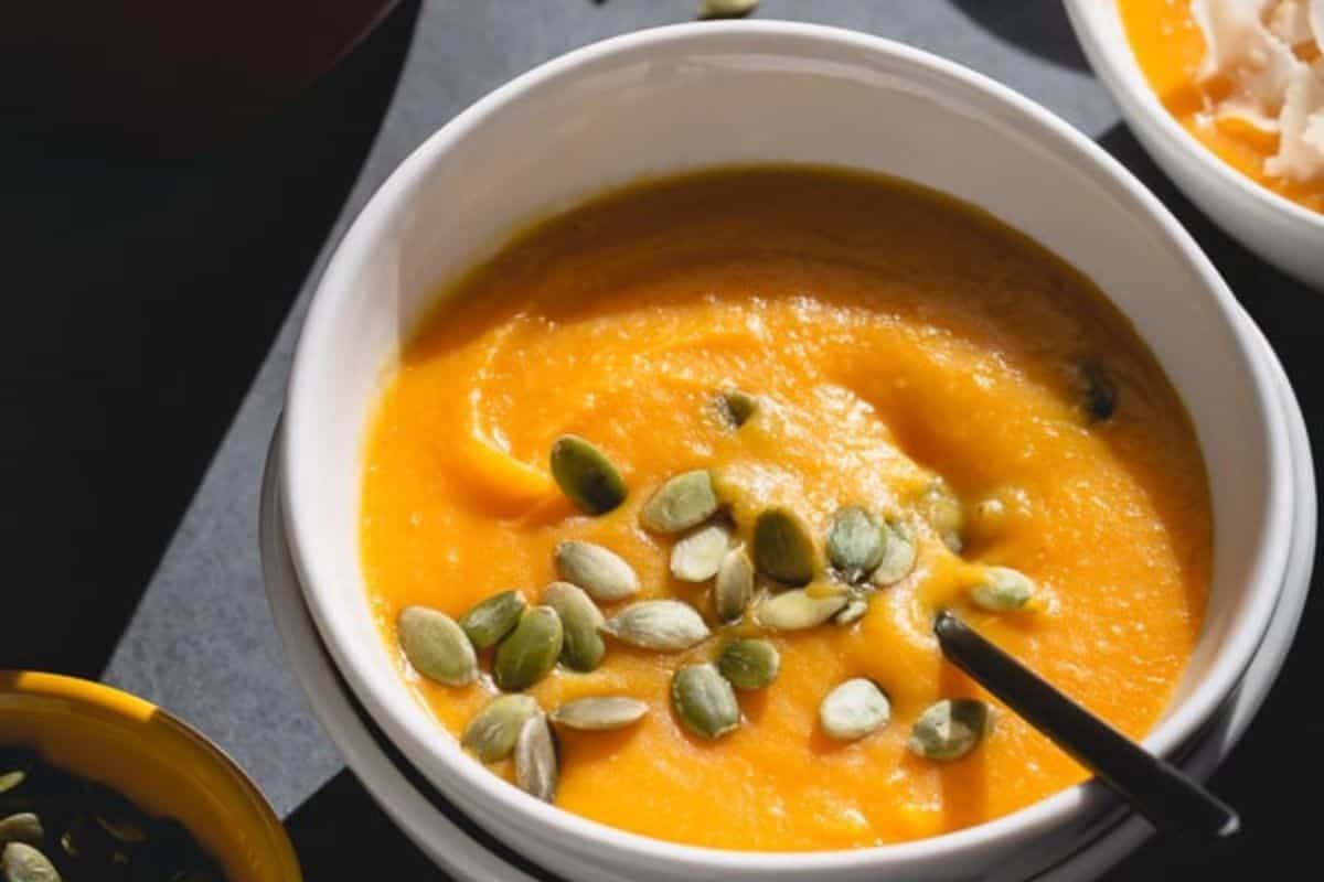 A bowl of orange soup, likely pumpkin or squash, topped with green pumpkin seeds and inspired by coconut recipes, with a spoon resting in the bowl.