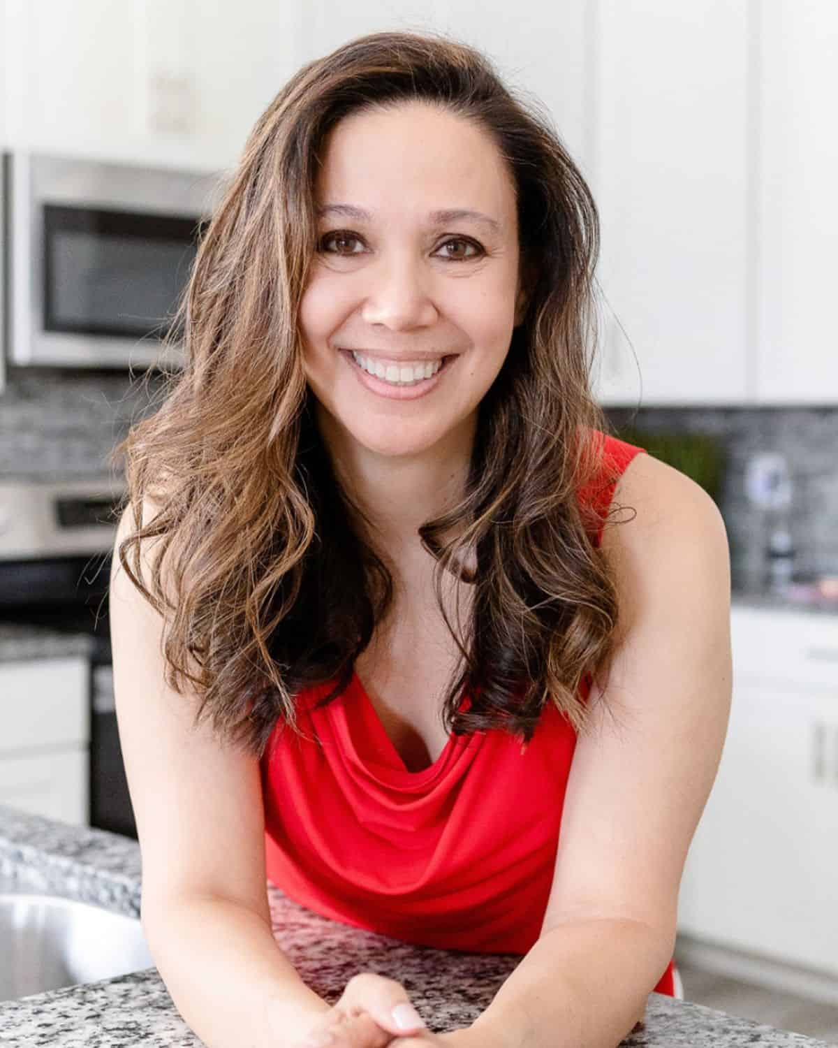 Corinne, with long brown hair and a red sleeveless top, smiles while leaning on a kitchen counter with white cabinets in the background, ready to share her Smart Meal Tips.