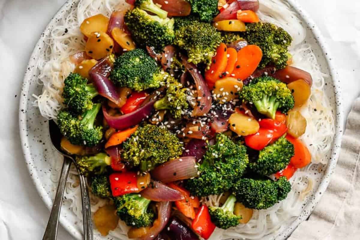 A plate of rice noodles topped with stir-fried broccoli, red onion, carrots, pineapple, and red bell pepper, garnished with sesame seeds—a delicious choice even for picky eaters or those searching for new broccoli recipes.