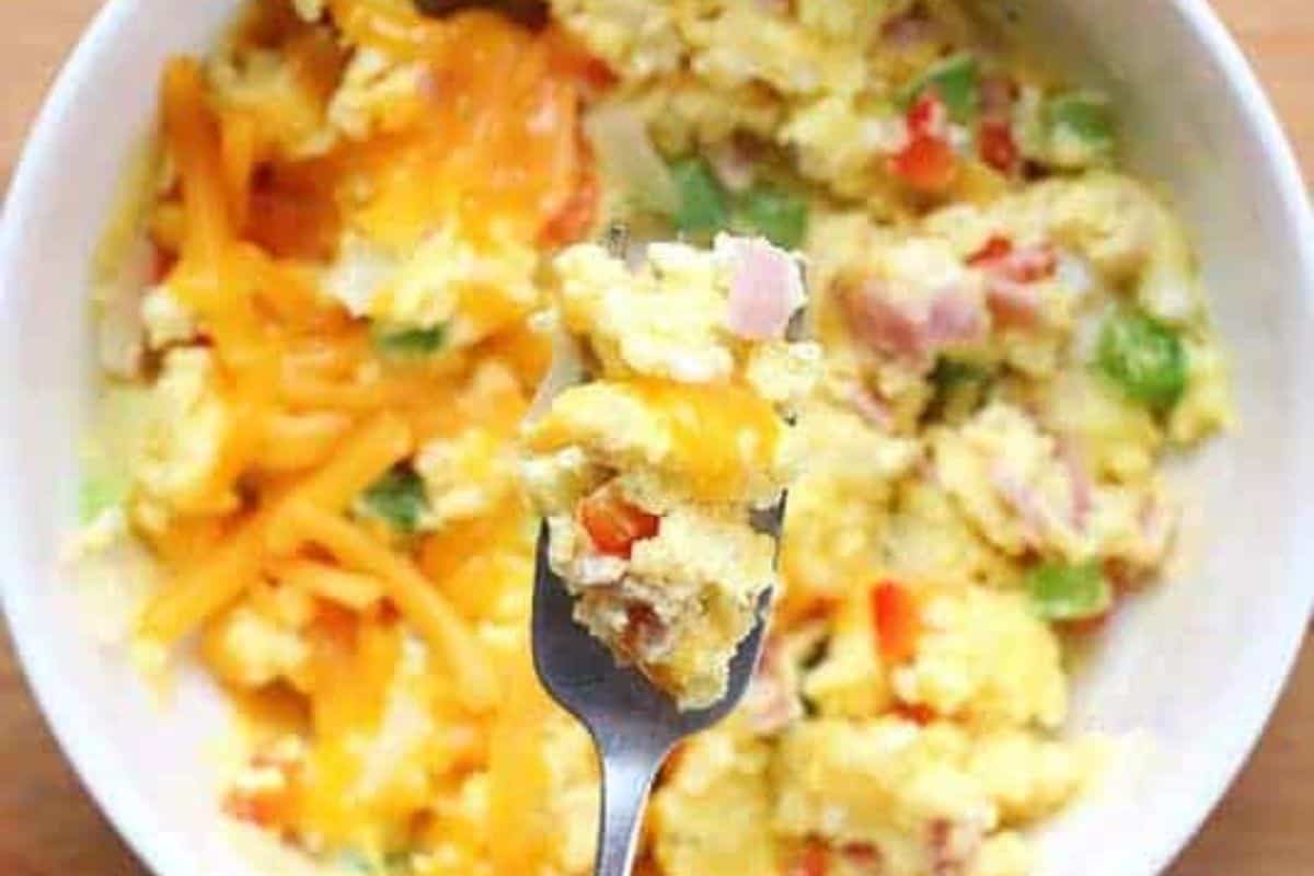 A close-up view of a bowl of scrambled eggs mixed with diced vegetables, melted cheese, and ham. The eggs are fluffy and golden, with visible pieces of red and green bell peppers and onions. A fork is lifting a portion of the scrambled eggs, showing the gooey melted cheese and colorful ingredients. The dish appears fresh and appetizing.