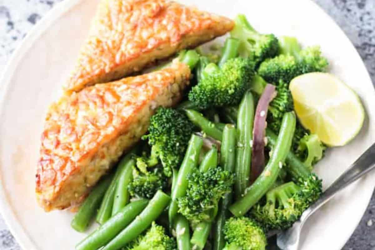 A plate with two triangular pieces of tempeh, steamed green beans, broccoli, red onion, a lime wedge, and a fork—perfect for picky eaters looking to try delicious broccoli recipes.