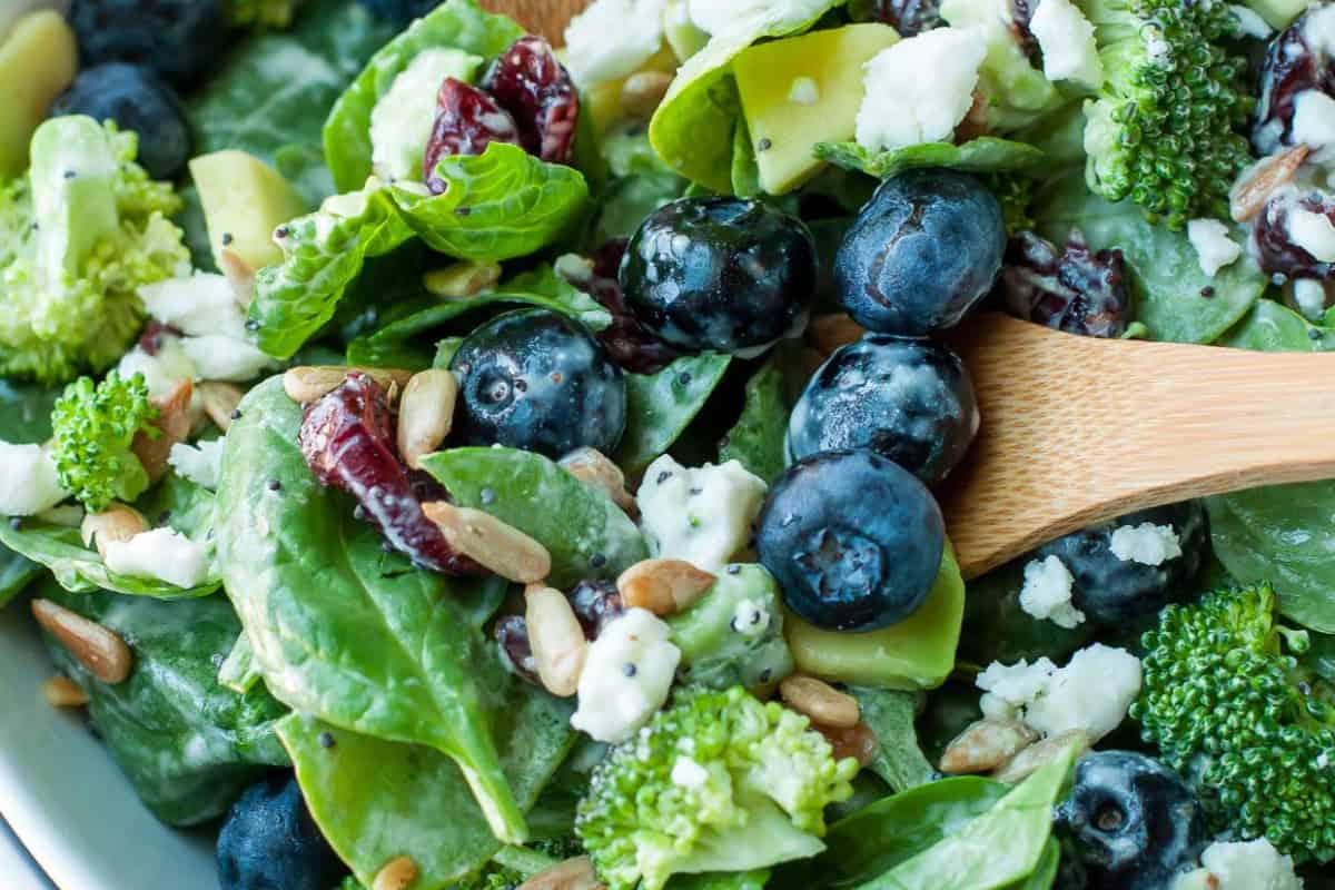 Close-up of a delicious salad with fresh spinach, blueberries, broccoli, sunflower seeds, dried cranberries, avocado, and crumbled cheese, mixed with a wooden spoon—perfect for picky eaters or as inspiration for new broccoli recipes.