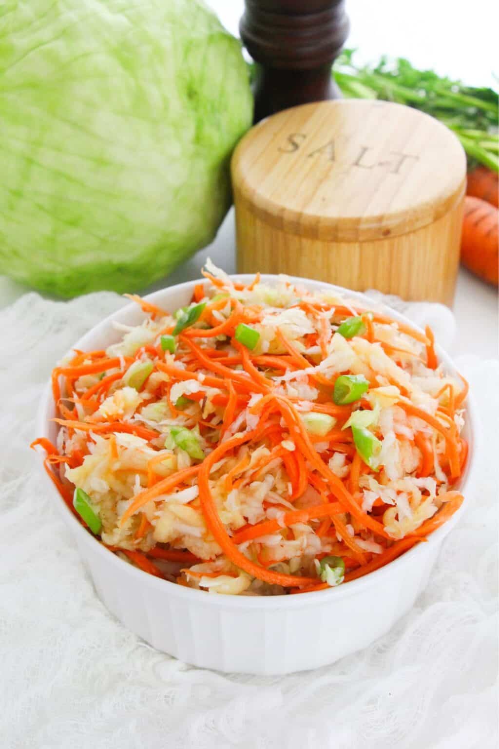 A white bowl filled with shredded cabbage and carrots sits on a white cloth, creating a fresh Carrot Slaw, with a cabbage, salt container, pepper mill, and carrot in the background.