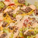 A close-up of a Big Mac Salad with ground beef, shredded cheddar cheese, lettuce, diced tomatoes, pickles, sesame seeds, and a creamy dressing drizzled on top.