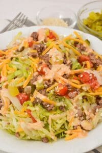 A plate of Big Mac Salad with ground beef, shredded cheddar cheese, lettuce, cherry tomatoes, and creamy dressing, accompanied by small bowls of pickles and sesame seeds in the background.