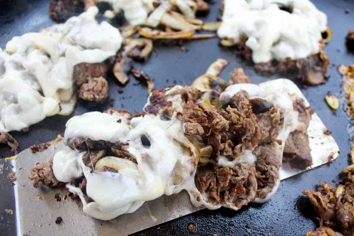 Cooked sliced beef, melted cheese, and sautéed onions on a Blackstone Griddle, with a metal spatula lifting one portion—perfect for flavor packed dinners and griddle recipes alike.