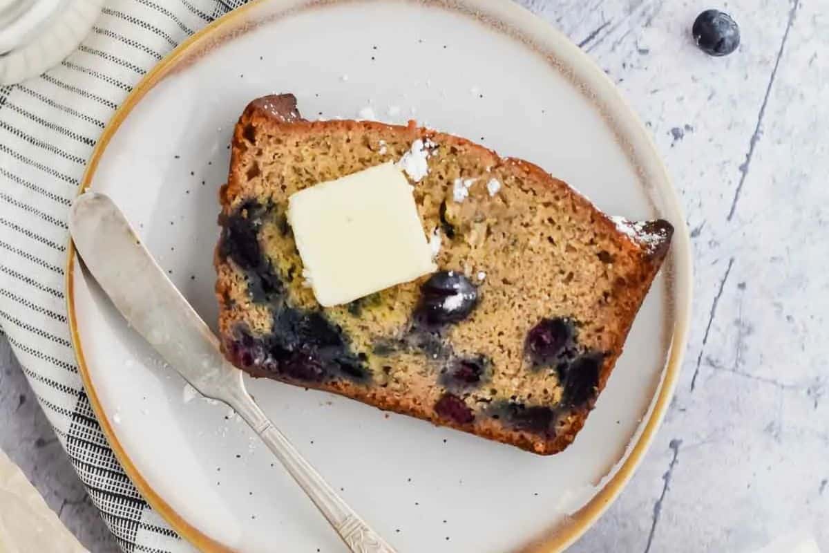 Close-up of a slice of blueberry banana bread with butter on top, served on a white and gold plate.