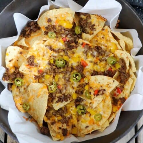 A cast iron pot filled with nachos topped with melted cheese, ground beef, chopped peppers, and sliced jalapeños, lined with parchment paper—a delicious choice for Dutch oven recipes and campfire cooking alike.
