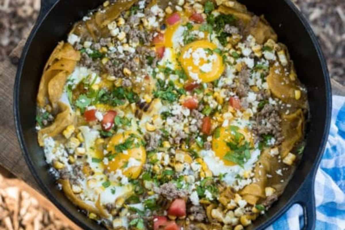 A skillet filled with chilaquiles topped with sunny-side-up eggs, ground meat, corn, tomatoes, cheese, and chopped cilantro—perfect for easy recipes or campfire cooking.