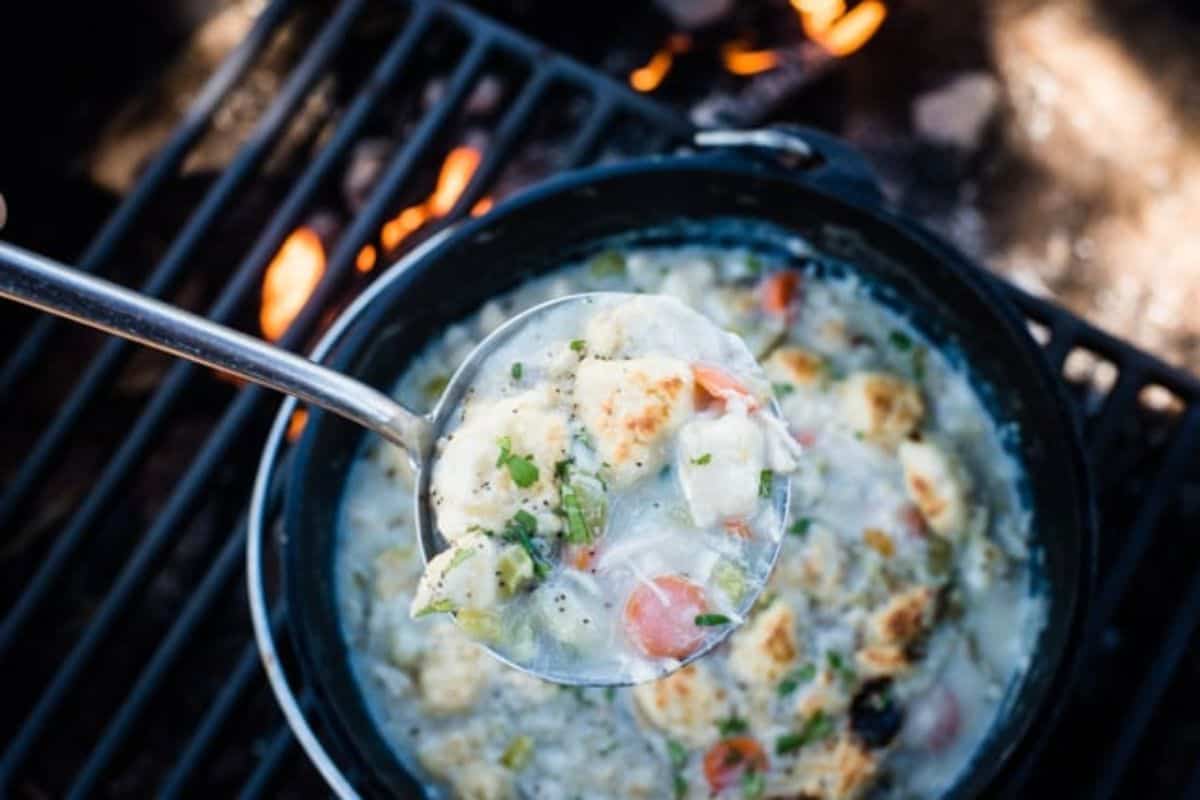 A ladle scoops a portion of creamy soup with vegetables and dumplings from a pot over a campfire grill—perfect for anyone who loves Dutch oven recipes and campfire cooking.