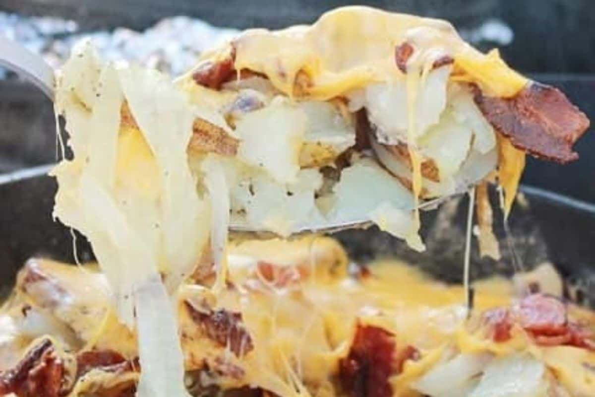 A close-up of a spoon lifting cheesy potatoes with bacon and onions from a pan, with melted cheese stretching—perfect for those who love easy recipes and Dutch oven recipes.