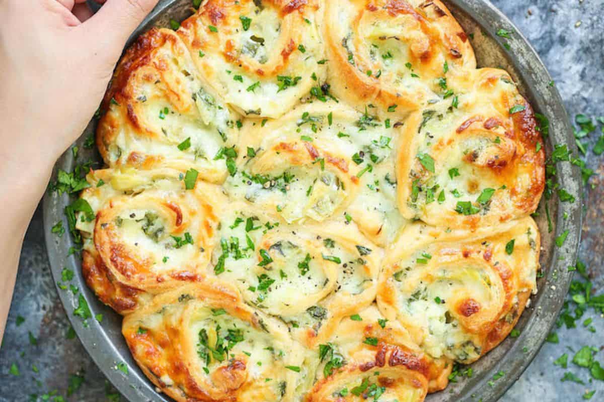 A round baking dish filled with golden, cheesy baked rolls topped with chopped herbs—an irresistible option for picky eaters seeking easy spinach recipes. A hand is holding the edge of the dish.