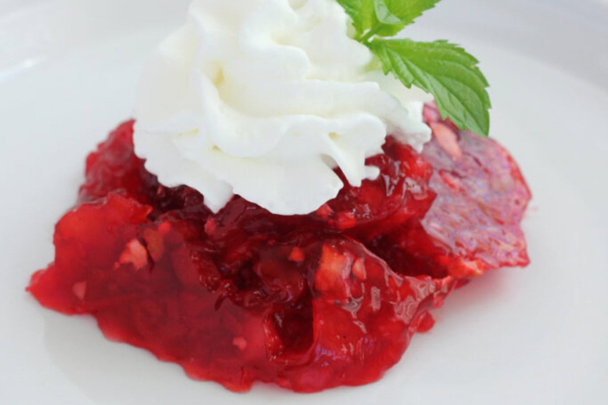 A serving of red gelatin dessert, inspired by Vintage Jello Salad Recipes, is topped with whipped cream and garnished with a sprig of mint on a white plate.