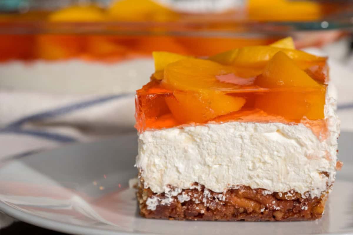 A square slice of dessert with a crumbly base, thick cream layer, orange gelatin, and peach slices on top sits on a white plate—reminiscent of vintage Jello salad recipes.