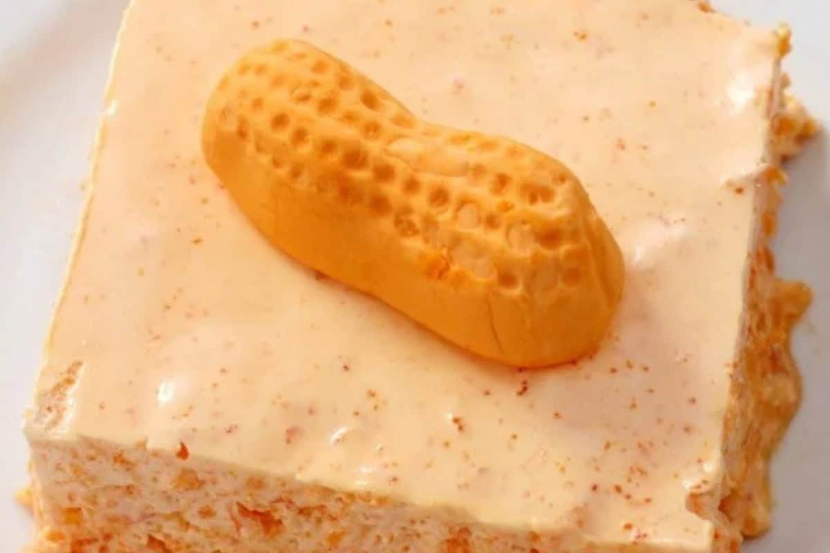 A square slice of orange peanut butter dessert, reminiscent of vintage jello salad recipes, topped with a single orange candy shaped like a peanut.