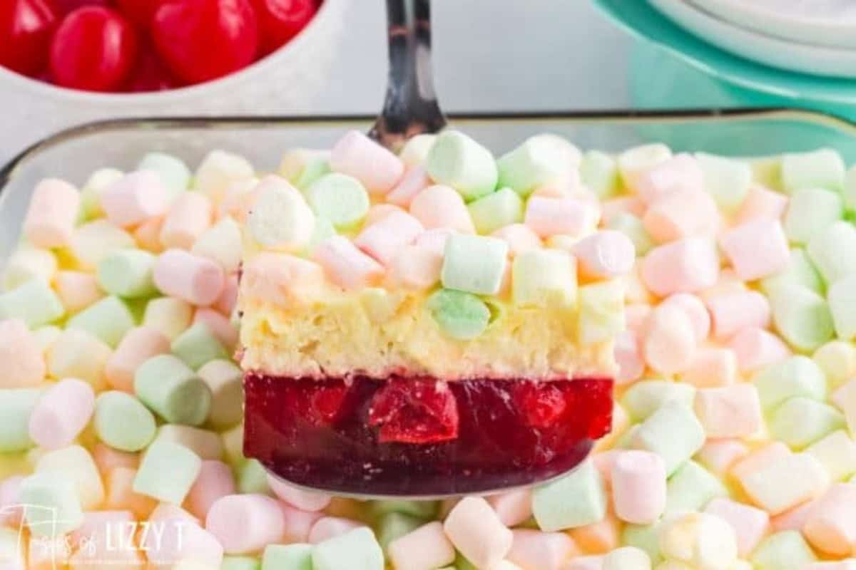 A dessert bar inspired by Vintage Jello Salad Recipes, featuring a cherry gelatin layer, creamy middle, and a topping of colorful mini marshmallows being lifted from the dish.