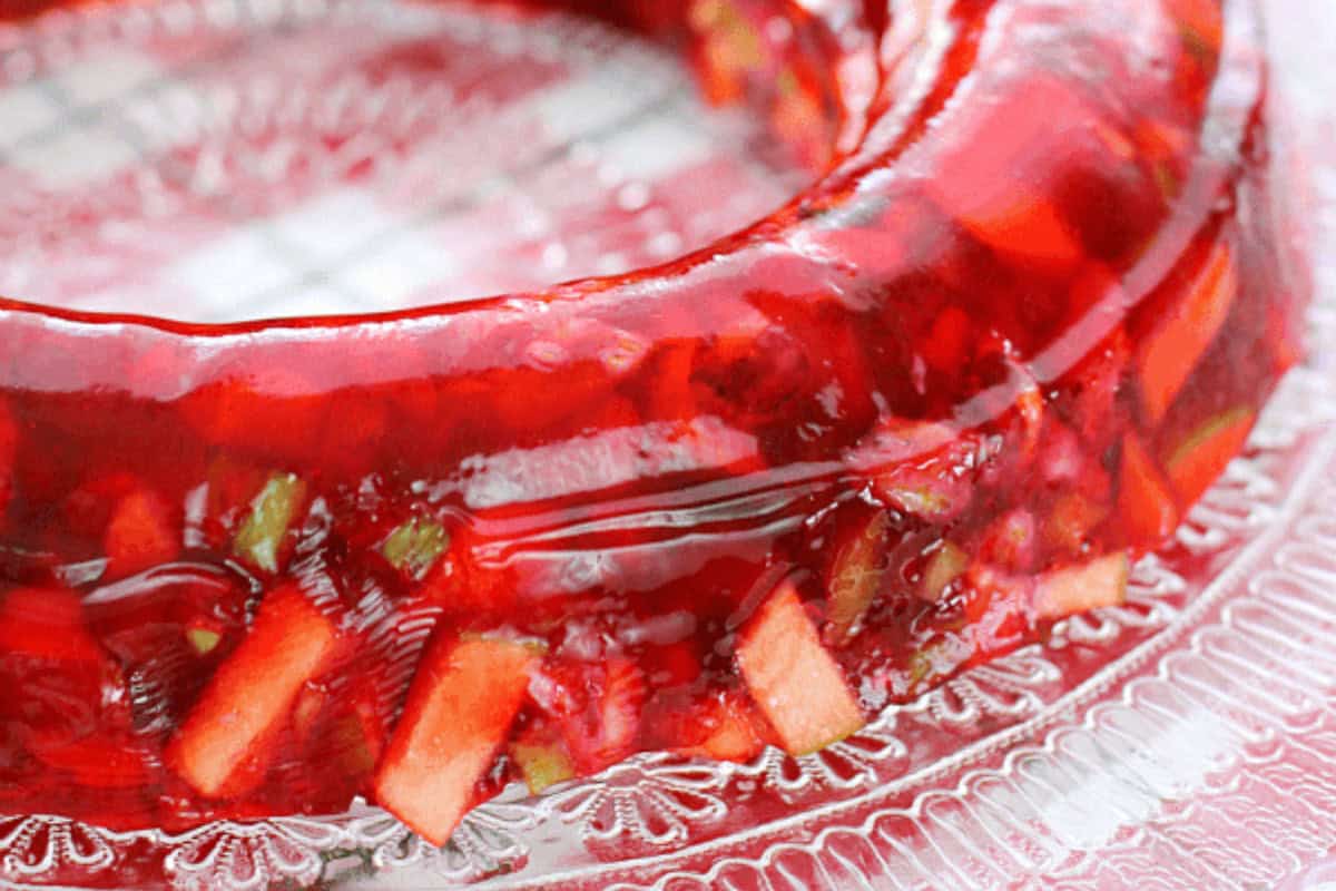 Red gelatin dessert mold with visible fruit pieces, displayed on a decorative clear glass plate—a classic nod to Vintage Jello Salad Recipes.