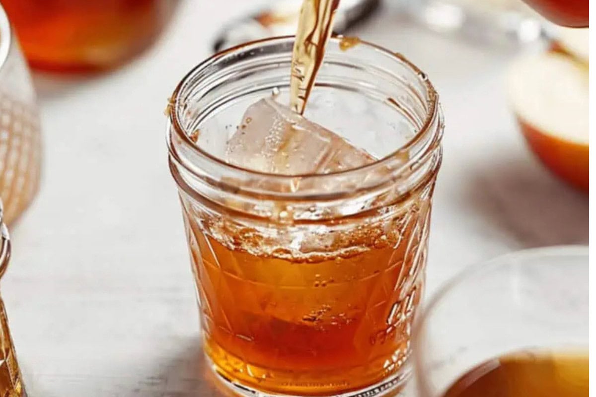 A close-up of a glass jar with ice cubes being filled with a brown liquid, possibly flavored moonshine or iced tea, on a light-colored surface—perfect inspiration for new flavored moonshine recipes.