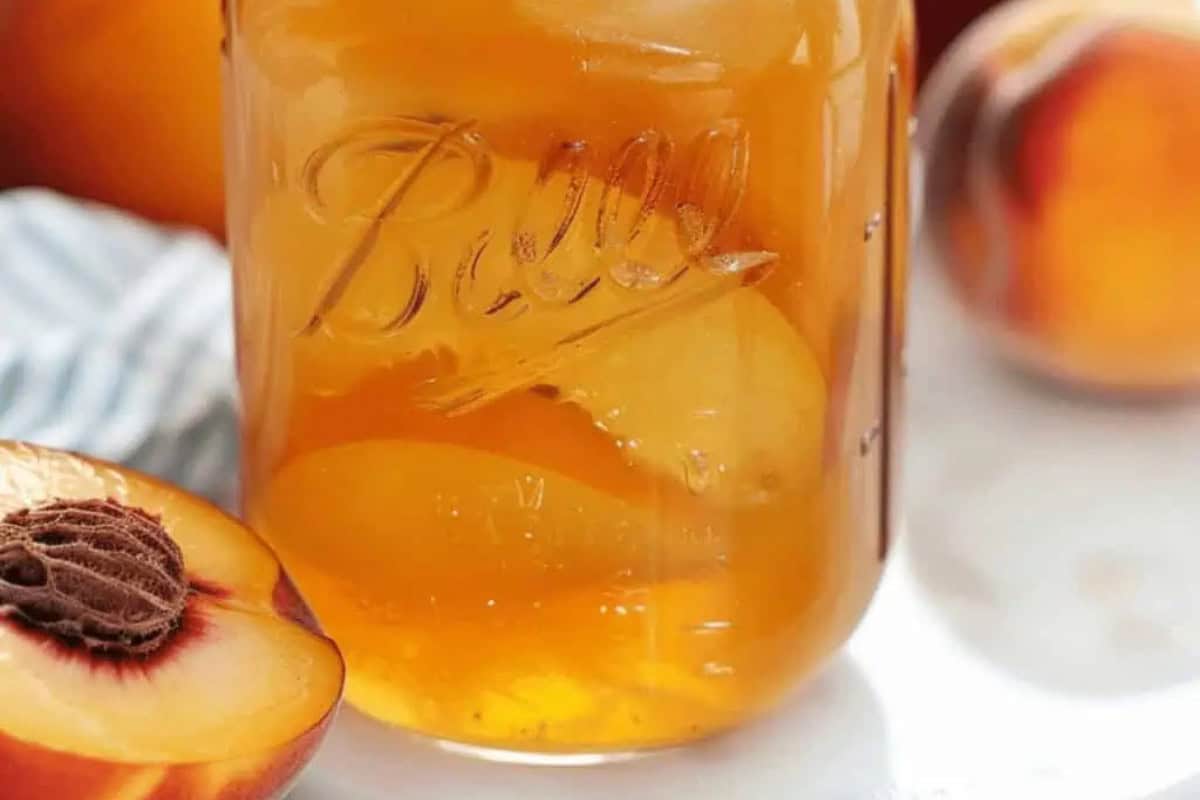 A glass jar filled with peach preserves sits next to a halved ripe peach on a white surface, perfect inspiration for must-try moonshine recipes.