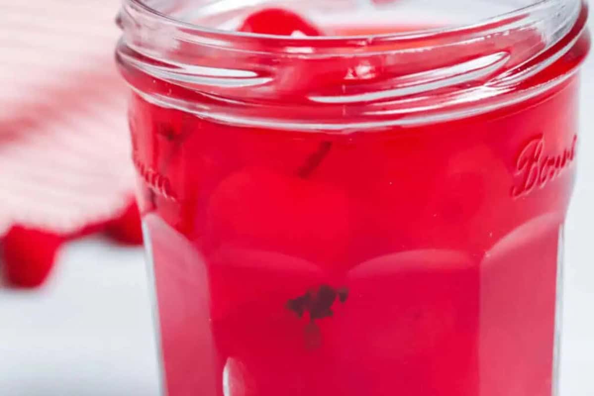 A glass jar filled with bright red maraschino cherries and syrup, perfect for creating must-try moonshine recipes, with a striped cloth partially visible in the background.
