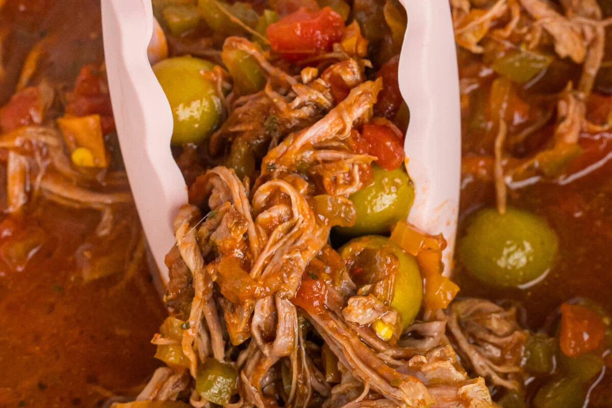 Close-up of shredded beef in tomato sauce with green olives and vegetables, being lifted by white tongs—a classic Cuban dish to spice up your next dinner.