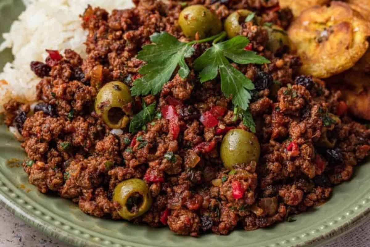 A classic Cuban dish, this plate of picadillo features ground beef, green olives, chopped vegetables, white rice, and fried plantains with a hint of spice, all garnished with a sprig of parsley for a flavorful dinner.