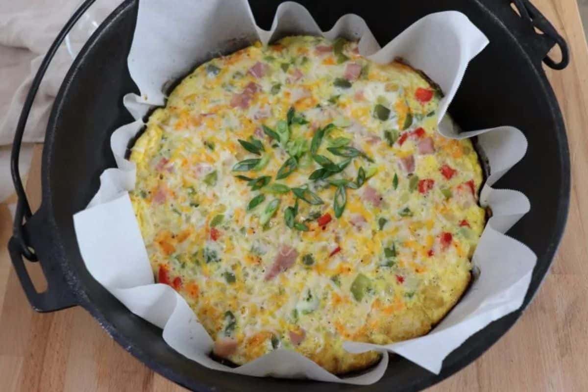 A baked frittata with chopped vegetables and ham, garnished with green onion slices, sits in a parchment-lined cast iron pan on a wooden surface—perfect for easy recipes and Dutch oven recipes alike.