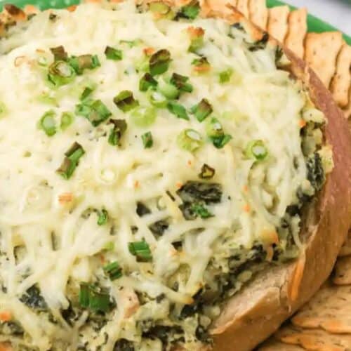 A round bread bowl filled with baked spinach artichoke dip, topped with melted cheese and green onions, surrounded by crackers on a green plate—a flavorful choice for easy spinach recipes and perfect for recipes for picky eaters.