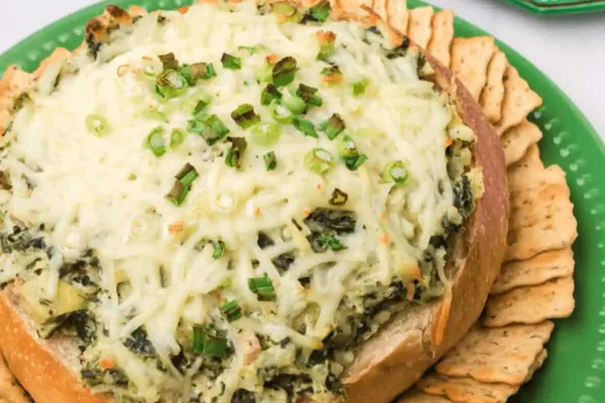 A round bread bowl filled with baked spinach artichoke dip, topped with melted cheese and green onions, surrounded by crackers on a green plate—a flavorful choice for easy spinach recipes and perfect for recipes for picky eaters.