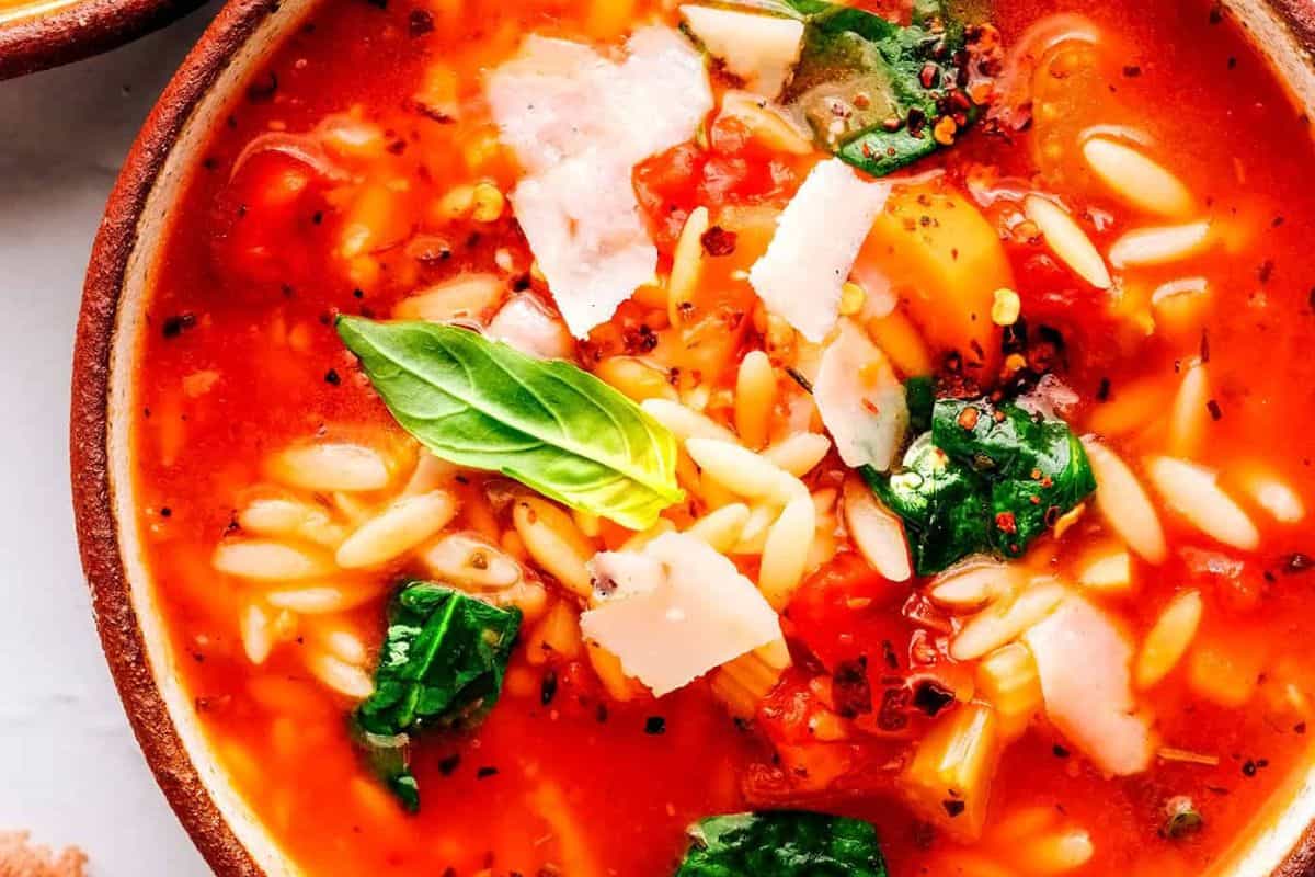 Close-up of a bowl of tomato-based soup with orzo pasta, spinach, basil, and shaved parmesan cheese—perfect for those seeking easy spinach recipes or delicious options for picky eaters.