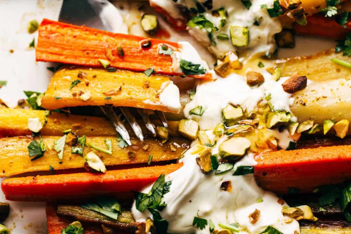 A close-up of roasted carrot sticks drizzled with a creamy white sauce and topped with chopped green herbs and nuts, with a fork resting among them.