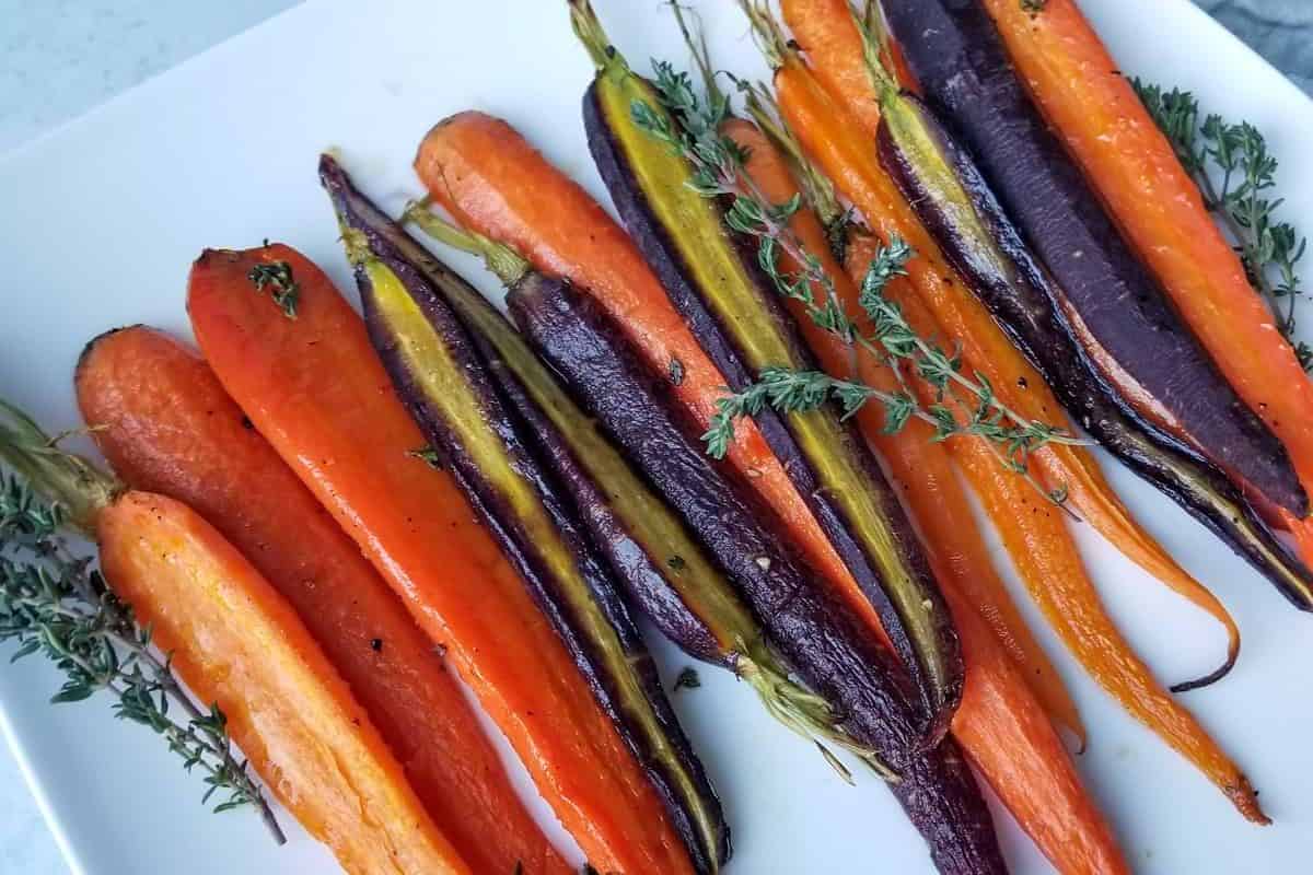 A white platter filled with a variety of roasted whole carrots in shades of orange, purple, and yellow, garnished with fresh thyme sprigs.