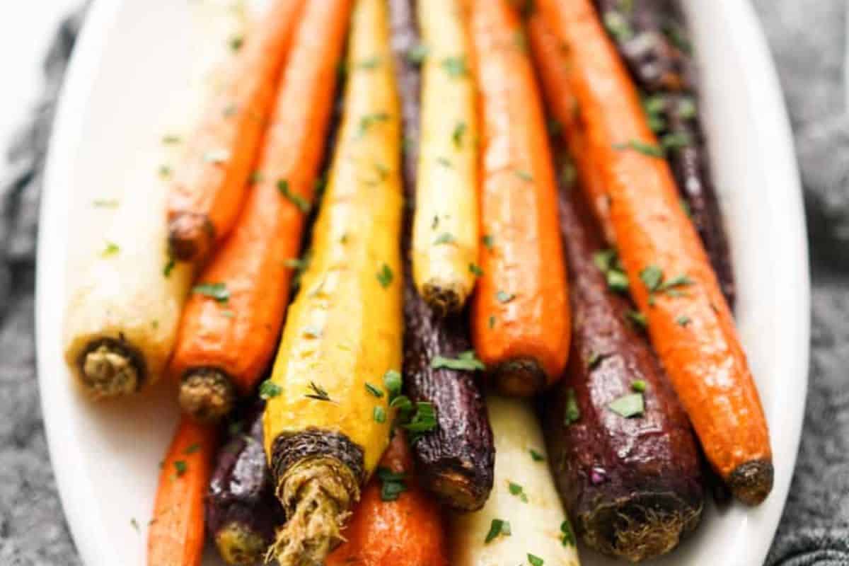 A white oval serving dish filled with an assortment of roasted whole carrots in shades of orange, yellow, white, and purple, garnished with fresh green herbs.