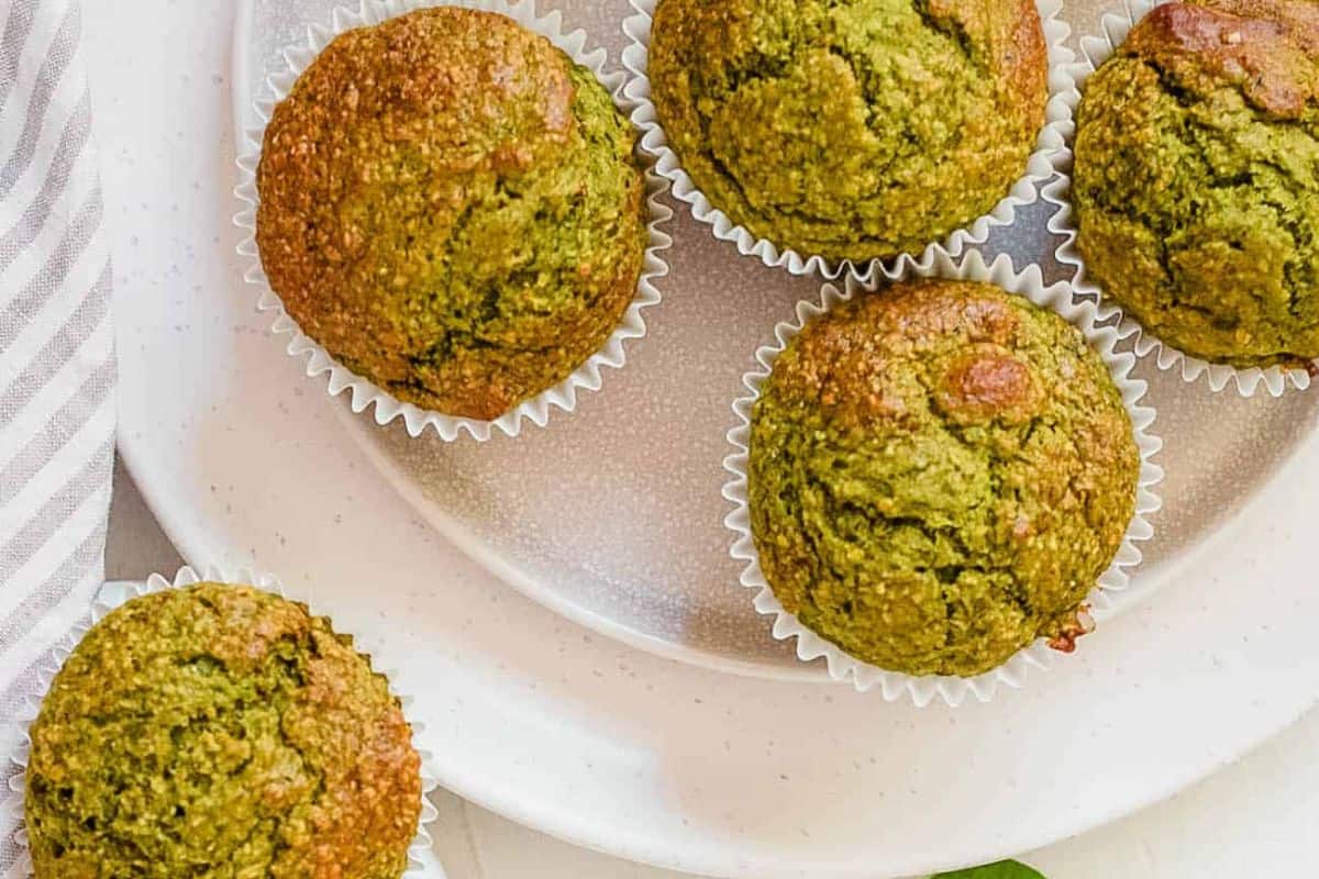 Five green muffins in white paper liners are arranged on a white plate next to a striped napkin—one of the easiest spinach recipes and perfect for picky eaters.