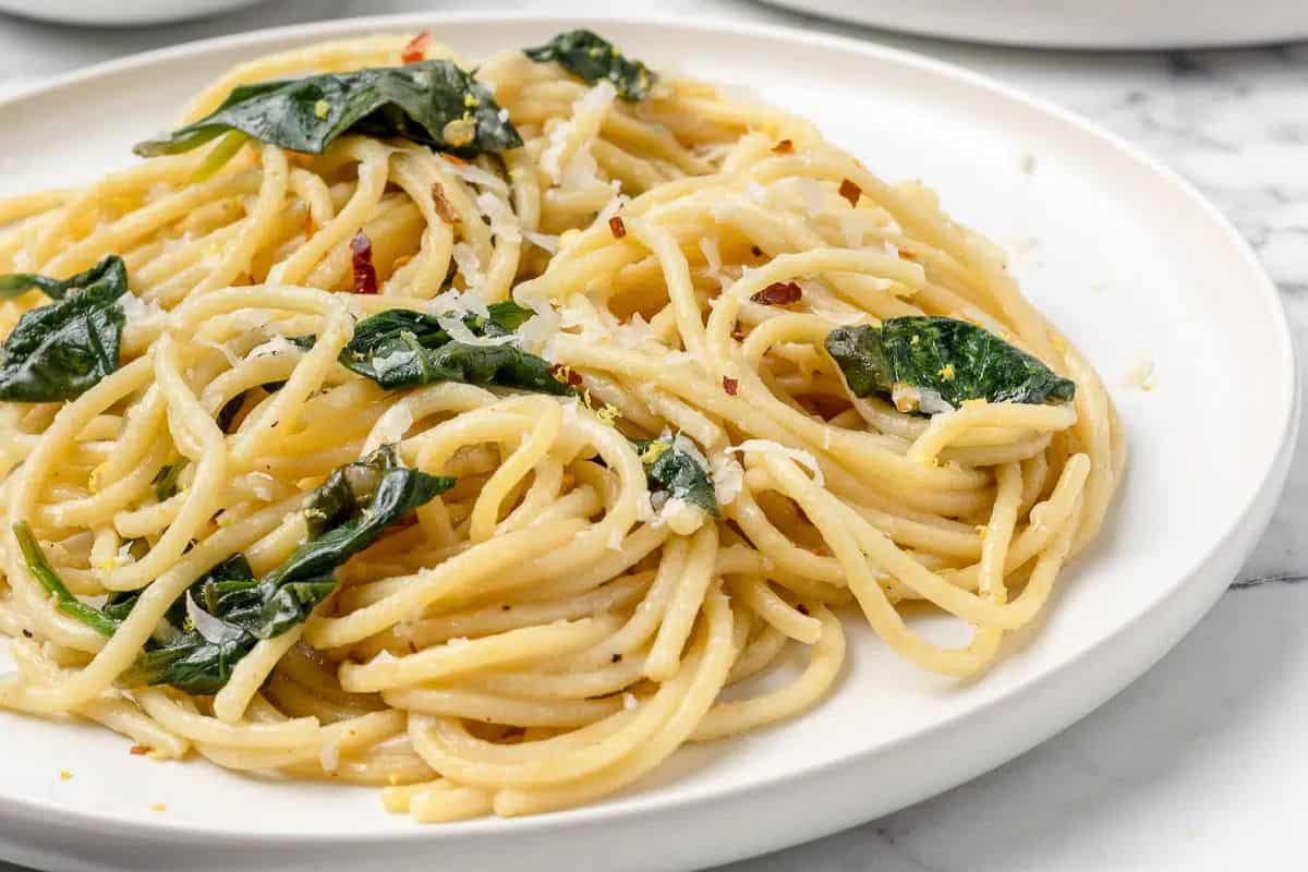 A white plate with cooked spaghetti mixed with wilted spinach leaves and grated cheese, on a marble surface—an easy spinach recipe perfect for picky eaters.