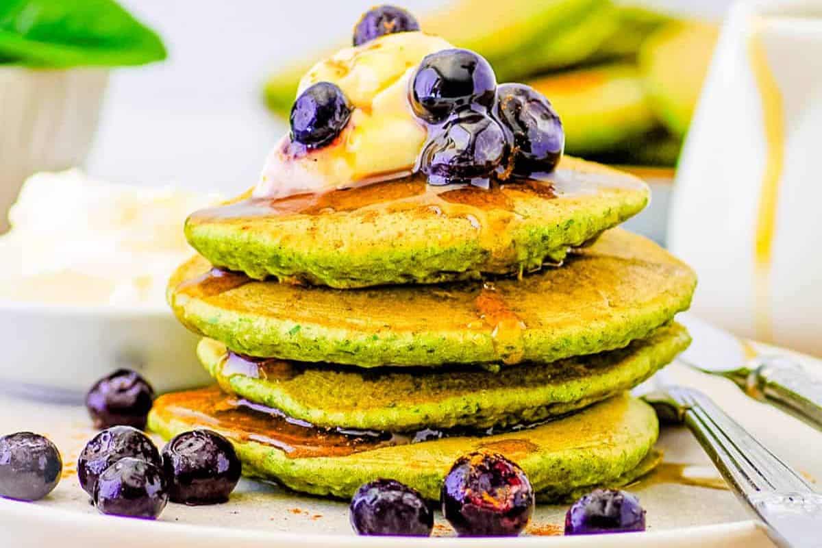 A stack of green pancakes made with easy spinach recipes, topped with blueberries, syrup, and a scoop of butter, served on a plate with extra blueberries and cutlery on the side.
