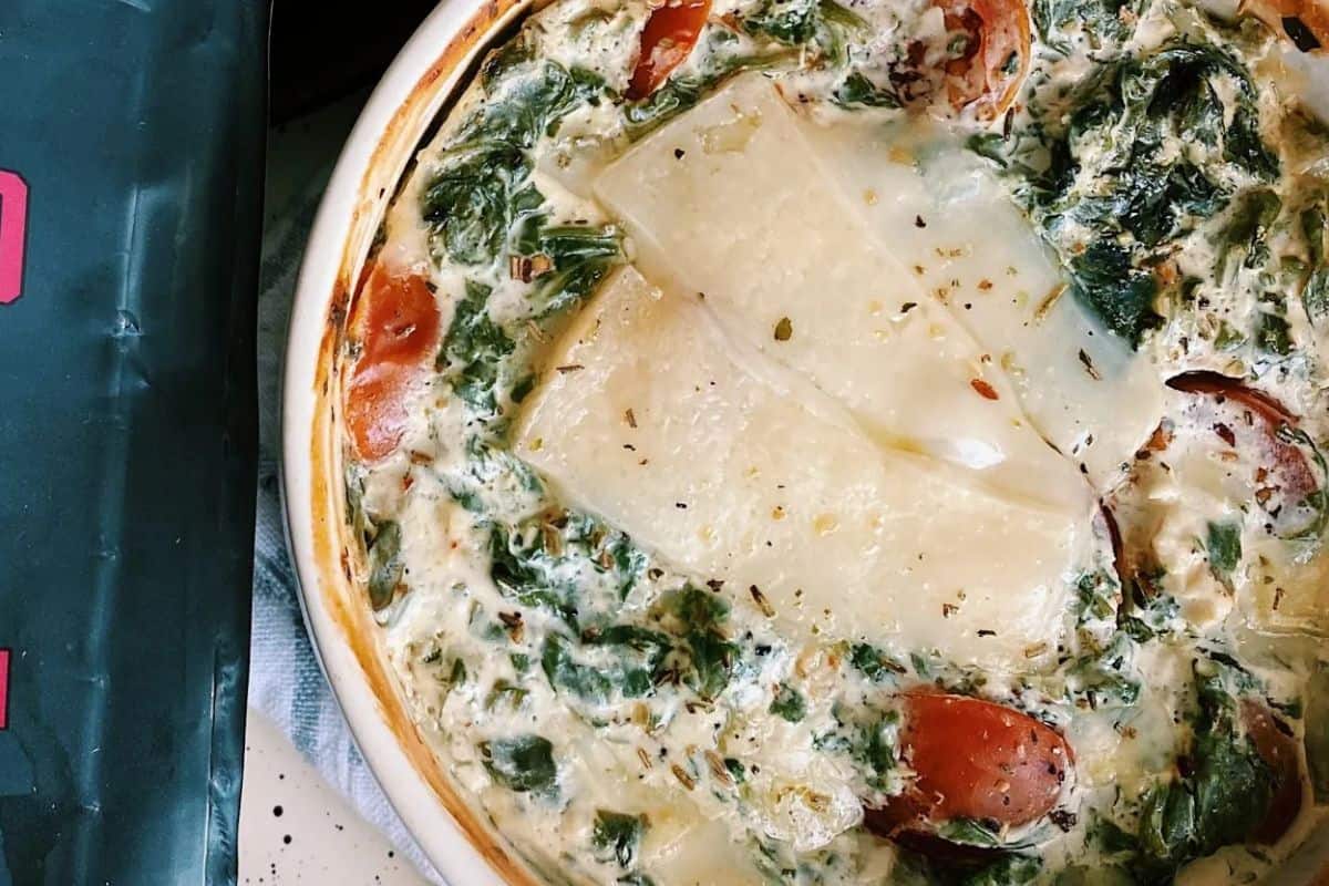 A baked spinach and tomato casserole topped with slices of cheese in a round white dish, perfect for picky eaters, shown next to part of a green package.
