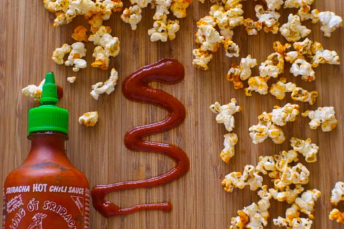 An overhead shot of a wooden surface with a bottle of Sriracha hot chili sauce, a zig-zag drizzle of Sriracha sauce, and scattered pieces of popcorn.