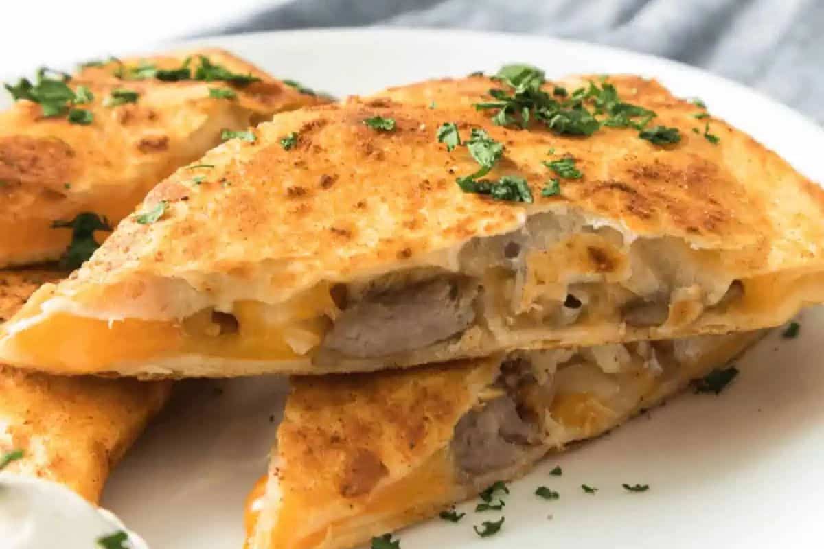 Golden brown steak quesadillas filled with melted cheese and steak slices, garnished with chopped herbs and served with a dollop of sour cream on a white plate.