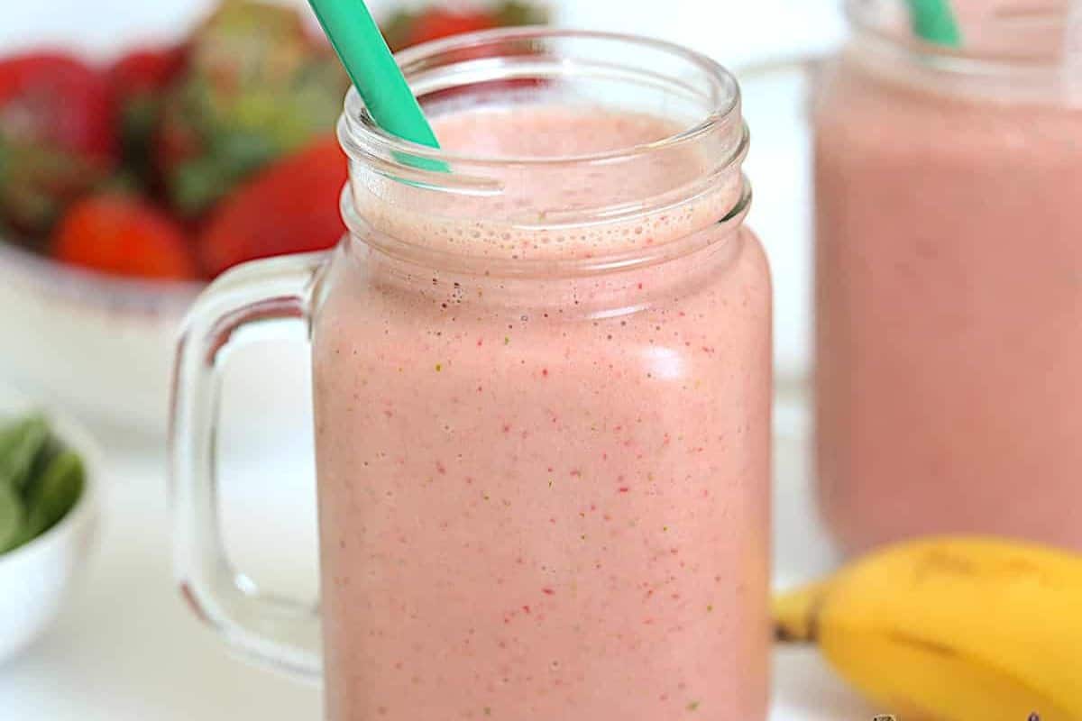 A mason jar filled with pink smoothie and a green straw, with strawberries and a banana in the background—perfect for picky eaters and great as one of your easy spinach recipes.