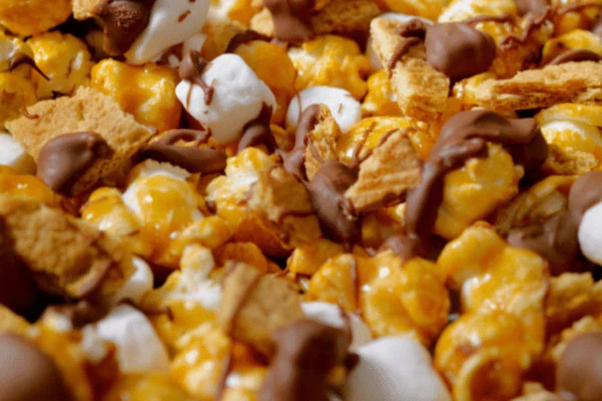 A close-up, overhead view of a sweet and salty s'mores popcorn mix, featuring caramel-coated popcorn, white marshmallows, graham cracker pieces, and chocolate chunks, with some elements drizzled in chocolate.