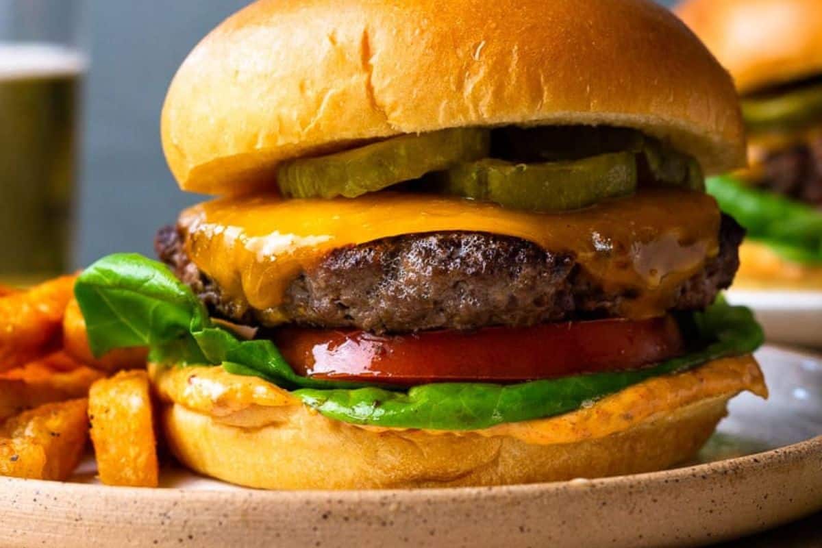 A savory venison burger with lettuce, tomato, pickles, cheddar cheese, and sauce in a bun, served with French fries on a plate—perfect for elevating your burger game.